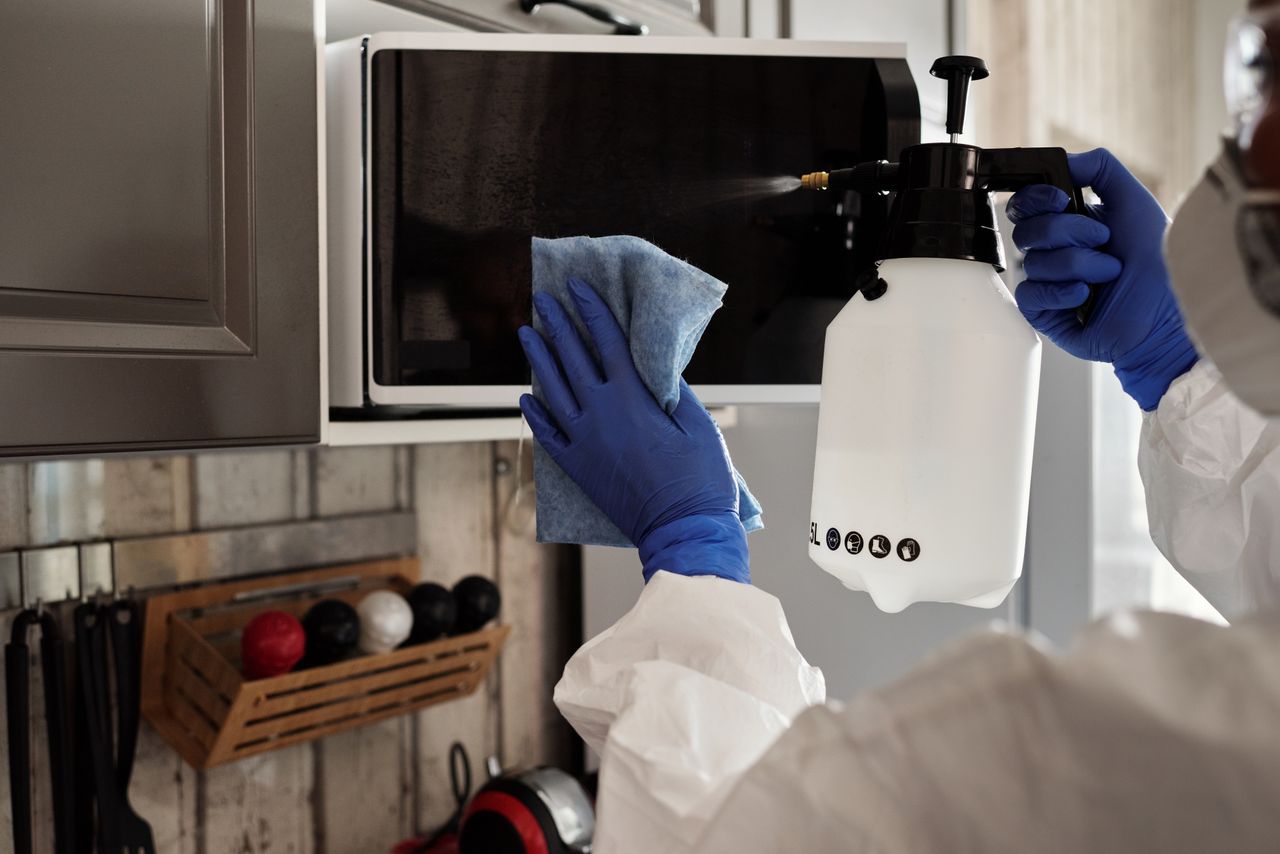 A person wearing protective equipment is cleaning a microwave indoors with a spray bottle and cloth.